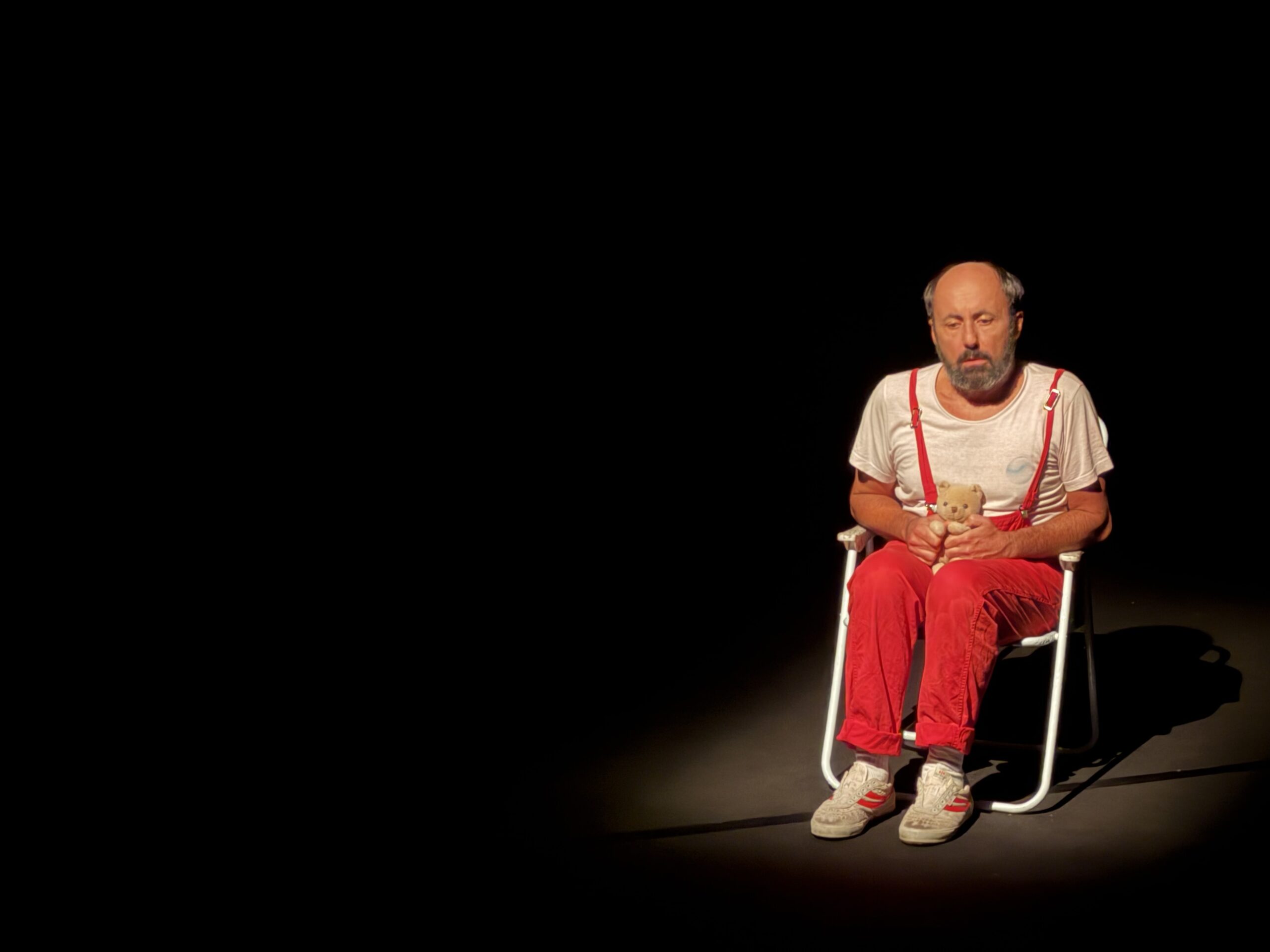 Lucio Patané sul palco recita seduto su una sedia. Indossa una tuta rossa e tiene in mano un piccolo orsacchiotto di peluche.