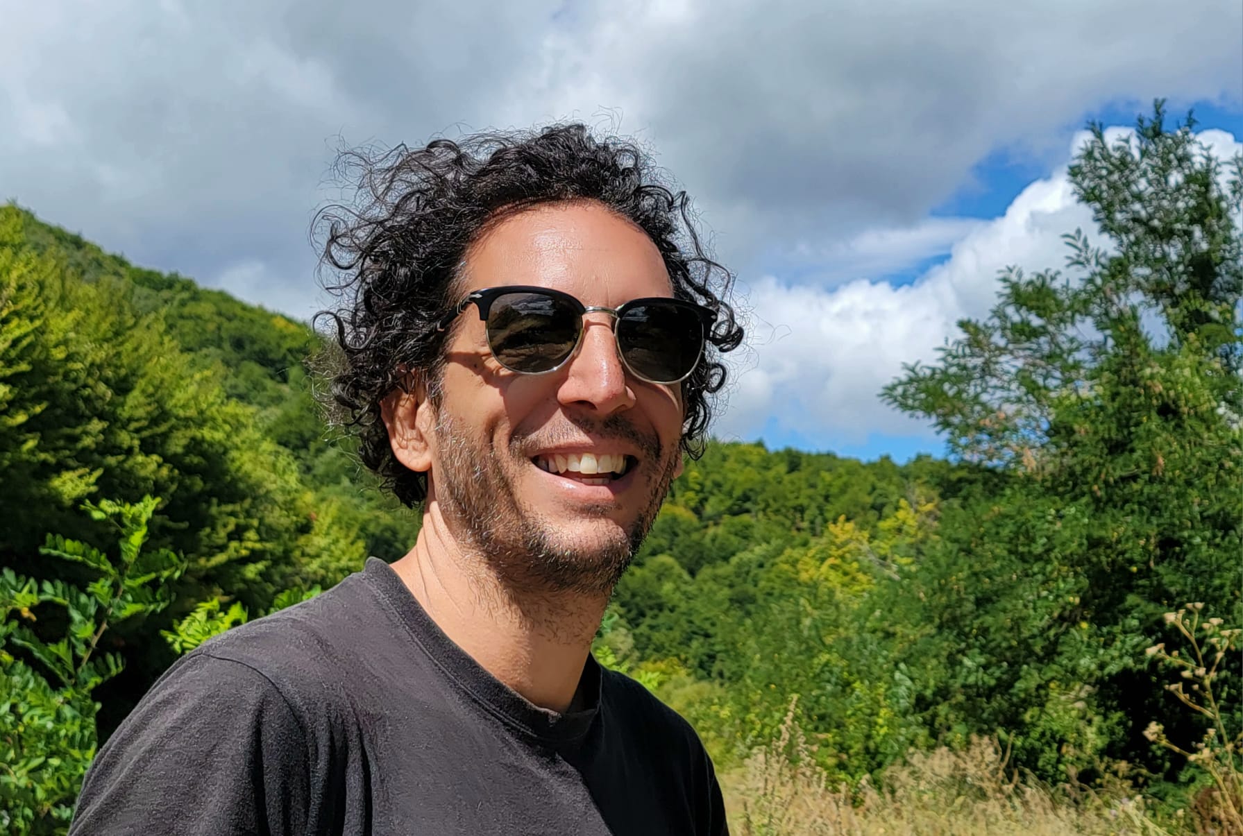 Stefano Usardi - regista cinematografico. Il regista è ripreso in primo piano con uno sfondo collinare alle spalle. È riccio e indossa occhiali da sole.