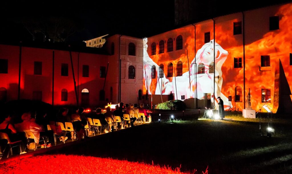 La macchina eroica e il percorso di gloria - esterno - in un ampio giardino siedono gli spettatori, Tasos Fotiou suona al sax, e su un muro è proiettata l'immagine de L'etasi di Santa Teresa