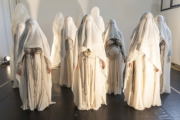 Il cast di Eumenidi in scena: tutte le attrici sono abbigliate con vesti bianche chiamate pepli e indossano il velo. Alcuni abiti hanno delle corde intorno ai seni o intorno a un pancione. Le interpreti sono disposte in più file.