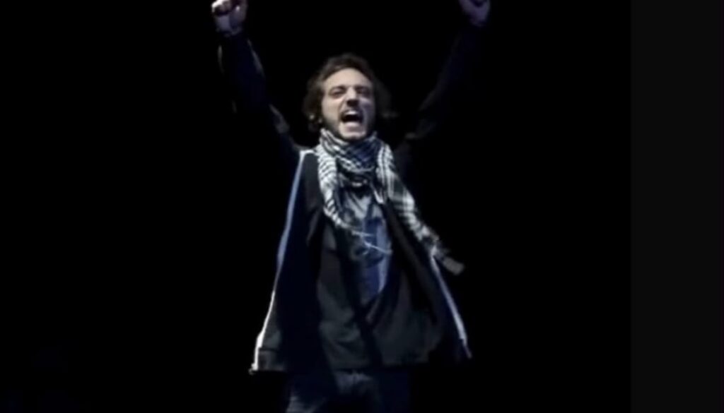 Uno dei protagonisti è in scena in penombra. Ha il braccio destro alzato, indossa una sciarpa a scacci bianca e nera. Il ragazzo grida.