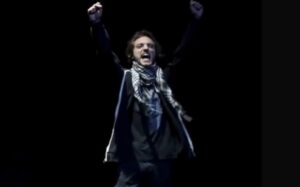 Uno dei protagonisti è in scena in penombra. Ha il braccio destro alzato, indossa una sciarpa a scacci bianca e nera. Il ragazzo grida.