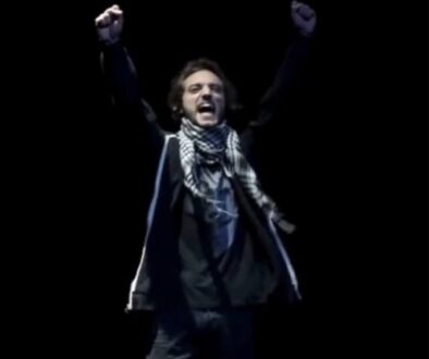 Uno dei protagonisti è in scena in penombra. Ha il braccio destro alzato, indossa una sciarpa a scacci bianca e nera. Il ragazzo grida.